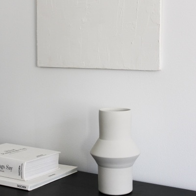 Vaso cerâmico branco com detalhes cinzentos numa superfície preta perto de livro branco e quadro abstrato branco.