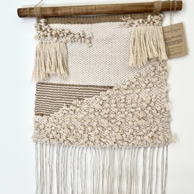 Tapeçaria pendurada em madeira com fios bege e castanhos e franjas.