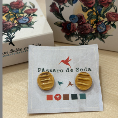 Brincos amarelo-mostarda redondos com textura apresentados numa cartolina com logótipo e caixas com ilustração floral