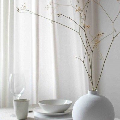Vaso arredondado branco com ramos secos e conjunto de cerâmica branca numa mesa com cortinas brancas