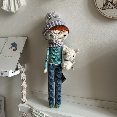 Boneco de crochê com gorro cinzento, cachecol, segurando ursinho branco numa prateleira com decoração de flâmulas de crochê