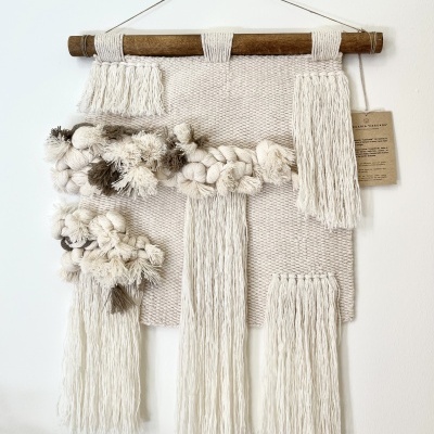 Tapeçaria decorativa em fios naturais bege e castanho clara com franjas e nós pendurada em ramo de madeira.