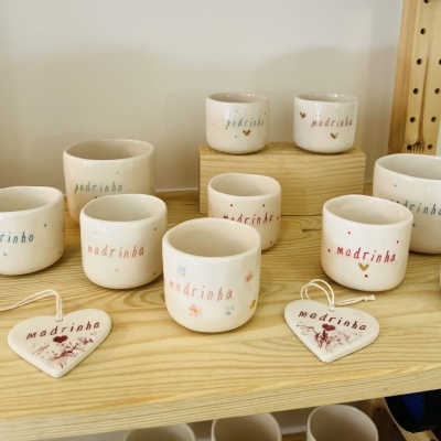 Pequenos vasos e corações em cerâmica com texto padrinho e madrinha em cores variadas, sobre prateleira de madeira.