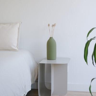 Mesa de cabeceira branca com vaso verde e flores secas ao lado de cama branca