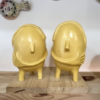 Estatuetas amarelas de cerâmica com braços cruzados sobre base de madeira e decoração colorida ao fundo