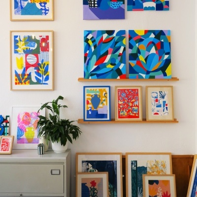 Vários quadros coloridos com padrões geométricos e florais emoldurados em madeira clara numa parede branca com planta e móvel cinza