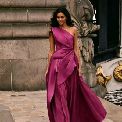 Vestido longo roxo assimétrico com um ombro, detalhes de pregas e folhos, ao ar livre perto de edifício de pedra com escultura.