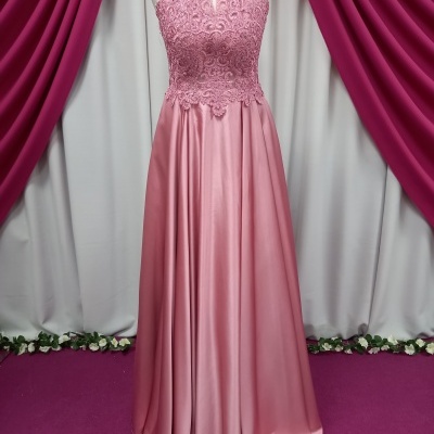 Vestido comprido cor-de-rosa com corpete em renda e saia lisa, colocado em manequim diante de cortinas cor-de-rosa e branco.