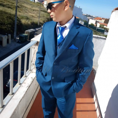 Rapaz jovem com fato azul e acessórios azuis numa varanda exterior.