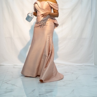 Mulher com vestido rosa claro satinada com ombros descubertos e mala prateada sobre chão marmorizado