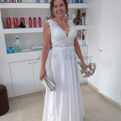 Mulher sorridente de vestido de noiva branco com renda segurando bouquet e mala numa sala com prateleiras e produtos.