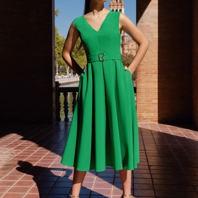 Mulher com vestido verde midi sem mangas e cinto, sandálias douradas, em fundo arquitetónico com azulejos e coluna.