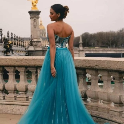 Vestido azul turquesa longo com corpete às riscas e cordões nas costas, em cenário urbano clássico.