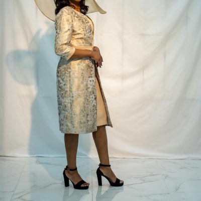 Mulher com vestido bege, casaco dourado estampado, sapatos pretos de salto e chapéu bege decorativo