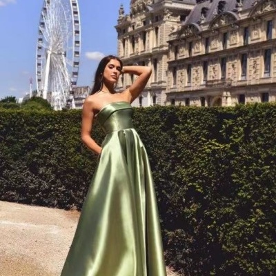 Mulher com vestido comprido verde esmeralda de cetim ao ar livre junto a jardim e prédio histórico.