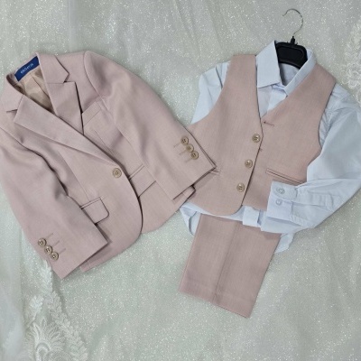 Conjunto infantil bege claro com blazer, colete, calças e camisa branca sobre tecido brilhante e renda branca.