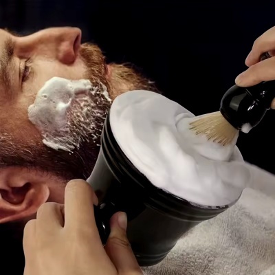 Homem a fazer a barba com espuma branca num recipiente preto e escova de barbear de cerdas claras