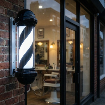 Lâmpada cilíndrica branca e preta estilo vara de barbear fixada numa parede de tijolos vermelhos junto a porta de vidro preto de barbearia.