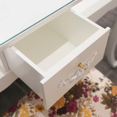 Gaveta branca aberta com puxador dourado em móvel branco com tampo de vidro sobre tecido floral colorido.