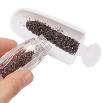 Frasco transparente despejando pequenas esferas metálicas prateadas numa bandeja branca de plástico