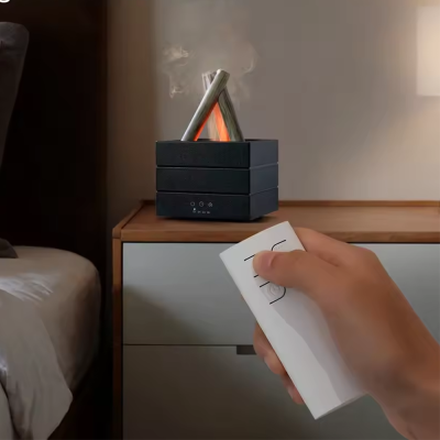 Humidificador preto a emitir vapor com forma quadrada, simulação de madeira em fogo, numa mesa de cabeceira ao lado de uma cama, mão segurando comando branco no primeiro plano.