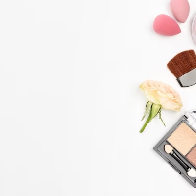 Produtos de maquilhagem variados sobre fundo branco, incluindo pó compacto, blush, verniz, sombra e pincel