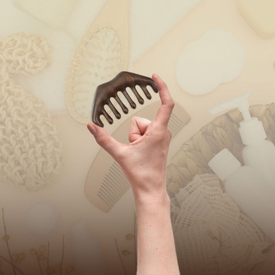 Pente de madeira escura com dentes largos segurado por uma mão com fundo de objetos de banheiro em tons bege