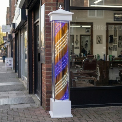 Coluna luminosa de barbeiro branco com LEDs azuis e brancos numa calçada à frente de barbearia