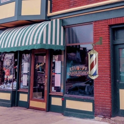 Fachada de barbearia com poste barbeiro listrado vermelho, branco e azul e toldo verde com riscas brancas