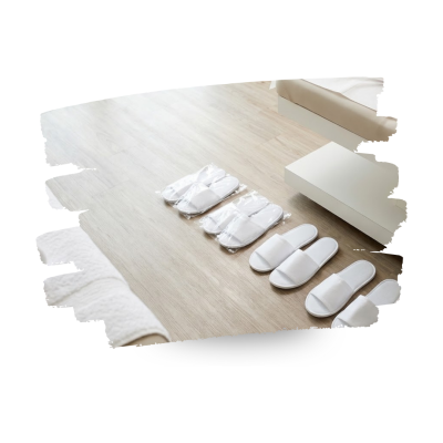 Quatro pares de chinelos brancos em chão de madeira clara ao lado de cama e tapete branco