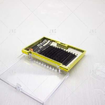 Estojo de pestanas negras com embalagem transparente e moldura dourada sobre superfície branca