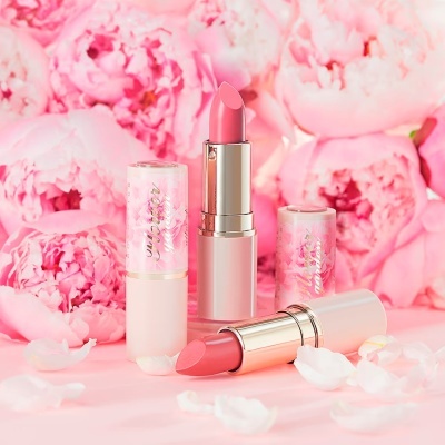 Dois batons cor-de-rosa com embalagens translúcidas decoradas com detalhes dourados sobre superfície rosa com pétalas e flores de peónias.