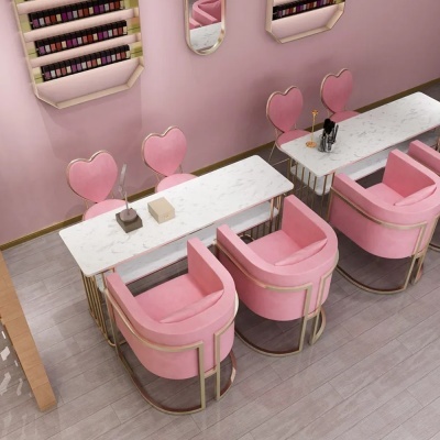 Espaço de salão com mesas brancas marmoreadas e cadeiras rosa com detalhes dourados, suportes de vernizes e espelhos ovais