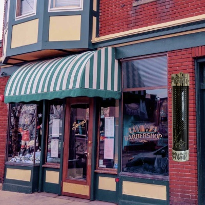 Fachada de barbearia com toldo às riscas verdes e brancas, porta de vidro, texto 'BARBERSHOP' na janela e parede de tijolos vermelhos.