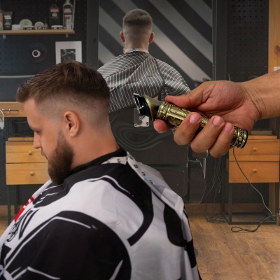 Máquina de cortar cabelo dourada perto da cabeça de homem com cabelo e barba, em salão com móveis de madeira.