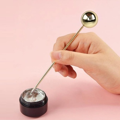 Mão a segurar bastão metálico dourado com esfera, a tocar creme prateado em frasco preto