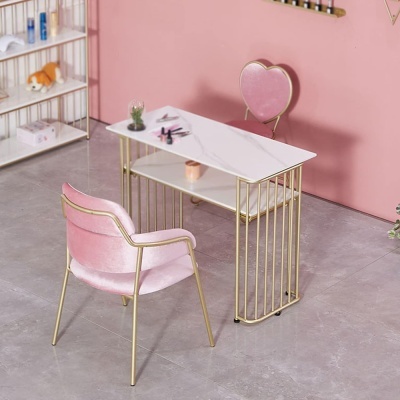 Mesa de estética branca com estrutura dourada, cadeira rosa de veludo e cadeira coração contra parede rosa