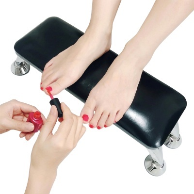 Suporte para pés com almofada preta e pernas cromadas, pessoa aplicando verniz vermelho nas unhas dos pés.