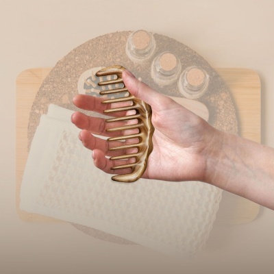 Pente dourado metálico segurado por mão sobre cortiça com frascos e pano branco