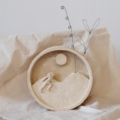 Escultura decorativa circular em tons bege com figura humana em relevo e arame com formas orgânicas.