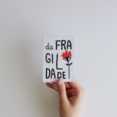 Cartão branco com a palavra 'da FRAGILDADE' e uma flor vermelha, segurado por uma mão.