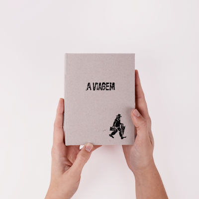 Livro cinzento com título A VIAGEM e ilustração preta sendo segurado por duas mãos