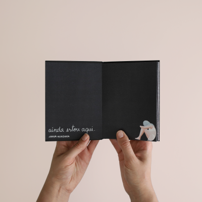 Livro aberto com texto e ilustração em páginas pretas, mãos seguram o livro, fundo bege