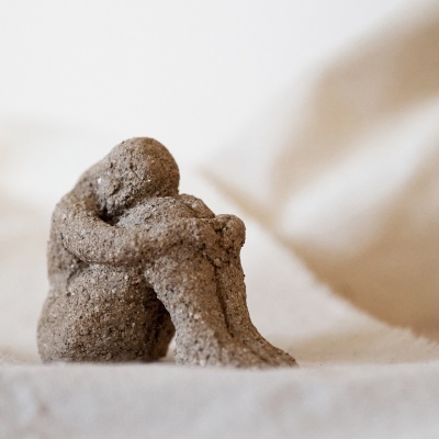 Escultura cinzenta texturizada de figura humana sentada abraçando os joelhos