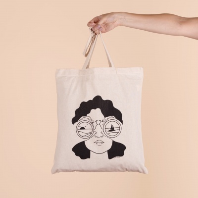 Mala de tecido cru com ilustração preta de rosto feminino com binóculos nas mãos.
