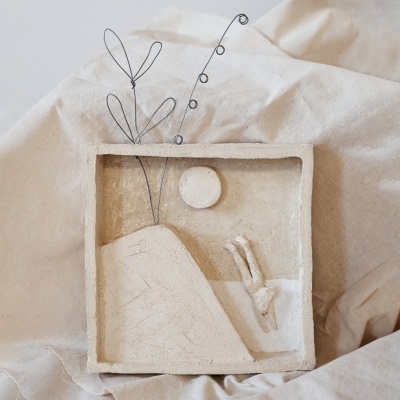 Escultura em relevo bege com figura a escorregar, círculo e plantas de fio metal