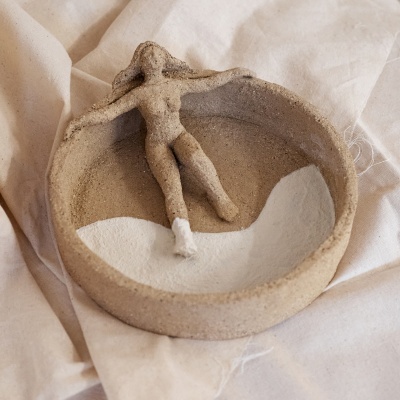 Prato decorativo de cerâmica bege com figura feminina reclinada e um pé branco