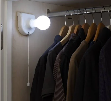 LÂMPADA PORTÁTIL LED INNOVA GOODS Lâmpada branca acesa numa base com cordão junto a varão com camisas escuras em cabides de madeira