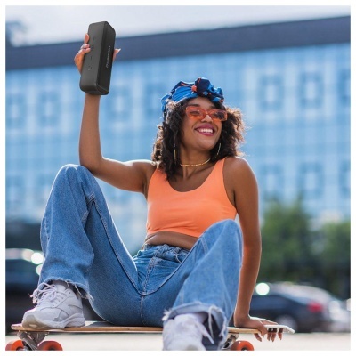 Mulher com top laranja e calças de ganga sentada numa prancha de skate a segurar uma coluna portátil preta