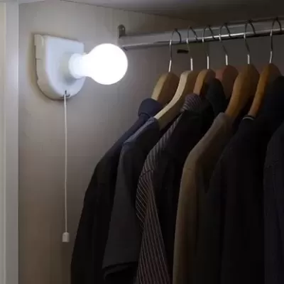 Lâmpada branca acesa numa base com cordão junto a varão com camisas escuras em cabides de madeira